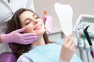 Woman with brown hair in dental chair looking into handheld mirror