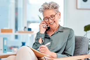 Woman in green shirt in chair writing and smiling while speaking on her cell phone