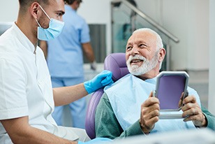 Man in dentist’s chair holding mirror looking at dentist who is wearing white scrubs