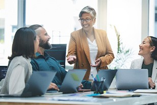 Woman with gray hair in tan jacket presenting to coworkers sitting around a table with laptops