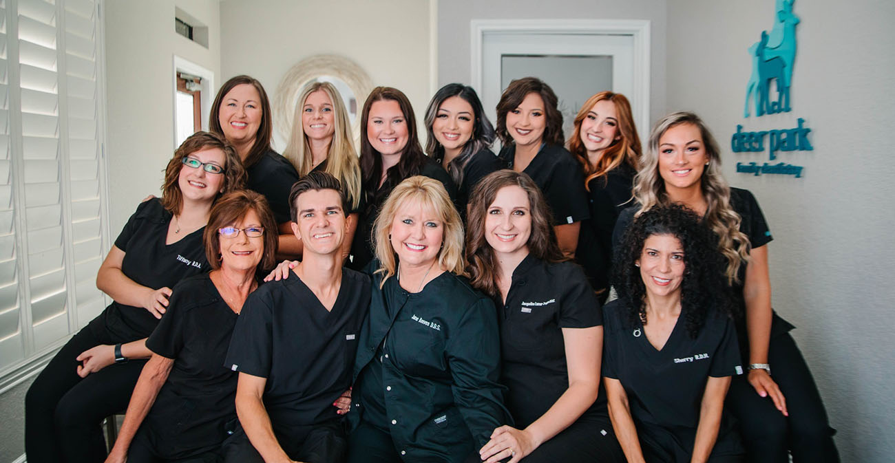 Smiling dental team group photo inside clinic – Deer Park Family Dentistry Group photo of the Deer Park Family Dentistry team, all dressed in black scrubs, smiling warmly while posing together inside the clinic with the practice’s logo visible on the wall – Deer Park Family Dentistry