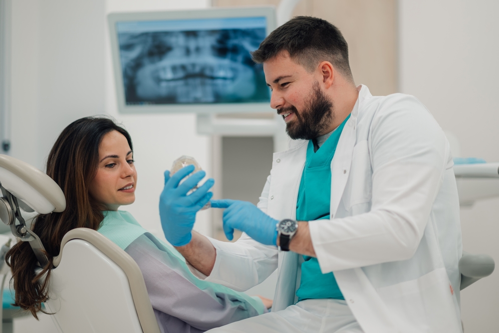 A dentist wearing gloves and a white coat shows a set of dentures to a female patient seated in a dental chair, explaining the procedure and fit while an X-ray image is visible in the background. - Dentures in Pasadena