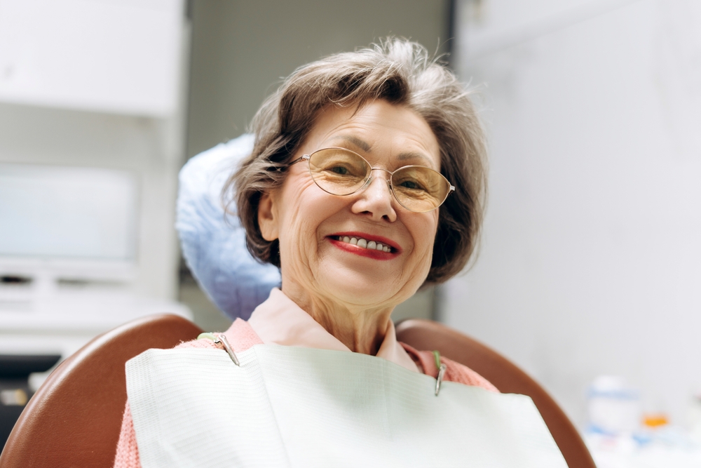 A smiling older woman wearing glasses sits comfortably in a dental chair with a protective bib, showcasing confidence and satisfaction with her dental visit.